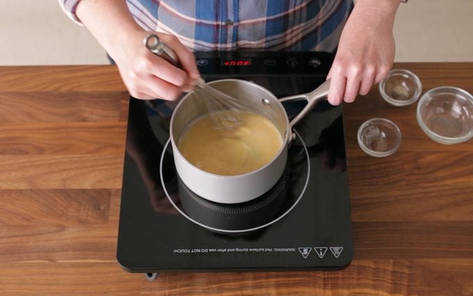 person Stirring a Blonde Roux with a whisk. roux pan is on a hot plate on a wooden countertop