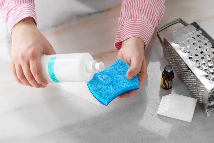 hand pouring Homemade Cleaners Dish Soap out of a squeeze bottle onto a sponge. bar of ivory soap and box grater nearby on the countertop