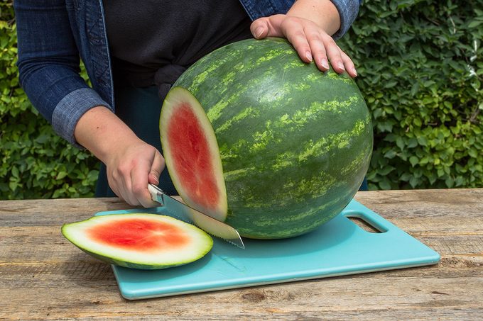 cutting the end off of a large watermelon on a blue cutting board on an outdoor table