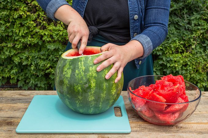 scooping watermelon out into a clear glass bowl on an outdoor table