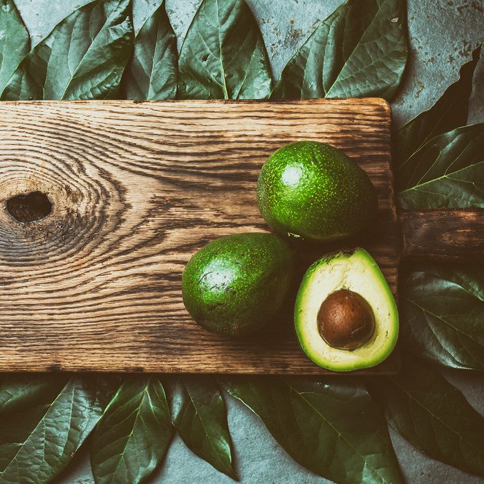 Food background with fresh avocado, avocado tree leaves and wooden cutting board.