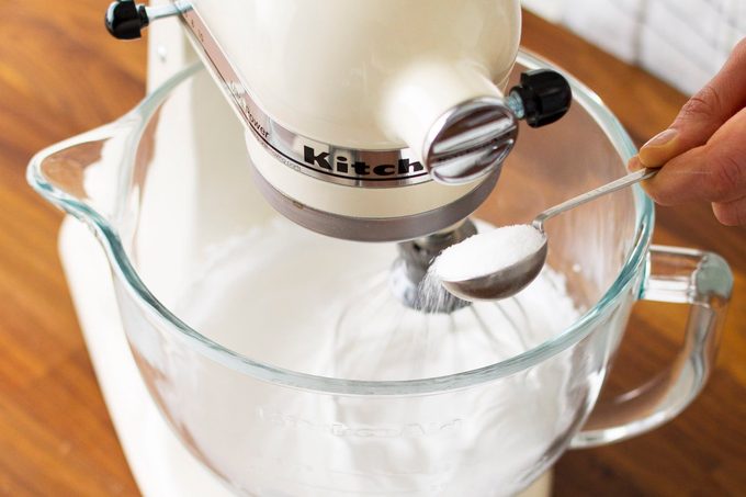A close-up of someone adding sugar to the bowl of a stand mixer as they make homemade meringue.