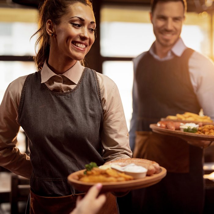 Two waiters serving lunch and brining food to their gusts in a tavern. Focus is on happy waitress. ; Shutterstock ID 1384574858; Job (TFH, TOH, RD, BNB, CWM, CM): Taste of Home
