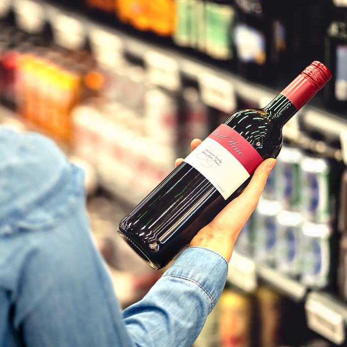 Woman reading the label of red wine bottle in liquor store or alcohol section of supermarket.