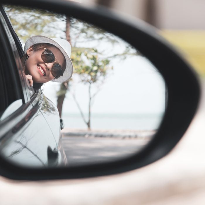 Asian woman in white shirt is looking into the side view mirror and smiling while sitting in her car, Travel concept.; Shutterstock ID 1407643370; Job (TFH, TOH, RD, BNB, CWM, CM): TOH Road Trip Games