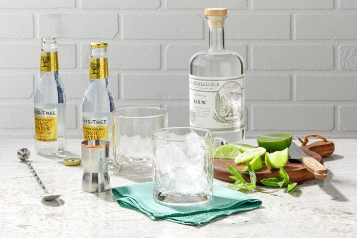 two glasses filled with ice surrounded by ingredients for a gin and tonic on a white countertop with a white brick background