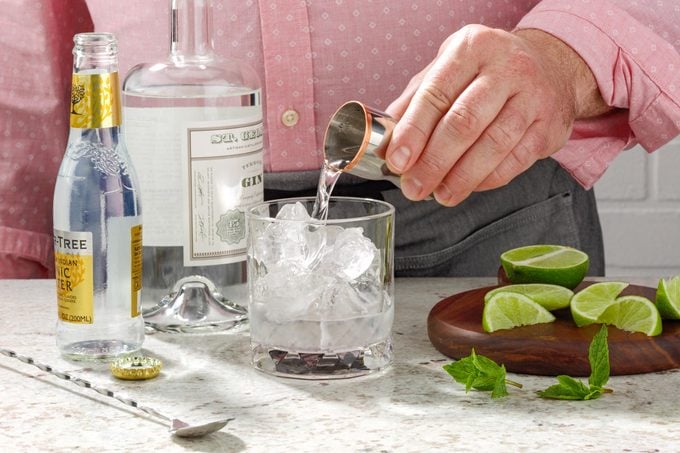 hands showing how to make a gin and tonic, pouring a shot glass of gin into a clear glass with ice on a marble countertop with limes and other bottles in the background