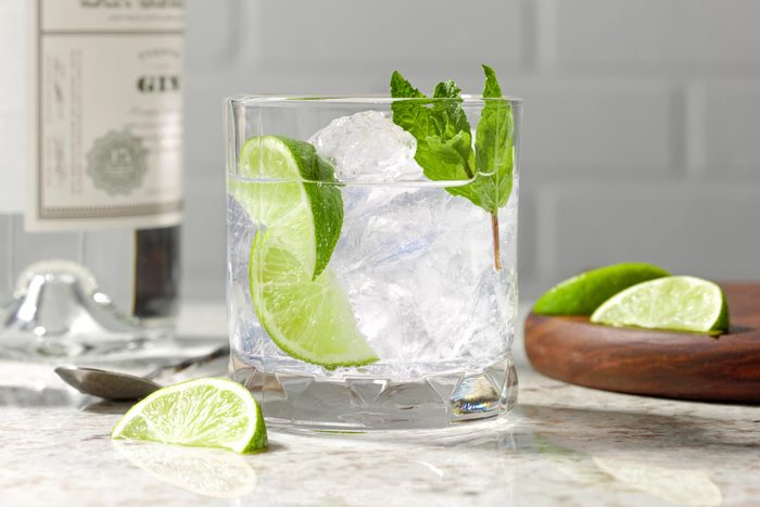 a clear glass with ice, clear cocktail, lime and herb garnish on a white marble counter top, with more limes and a bottle of gin in the background