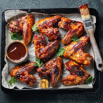 Roasted chicken wings in barbecue sauce with sesame seeds and parsley in a baking tray on a dark table. Top view. Tasty snack for beer on a dark background. Flat lay.