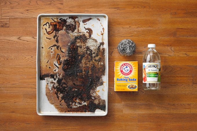 Dirty Baking Sheet next to box of baking soda, bottle of white vinegar, and steel wool on wood butcher block background
