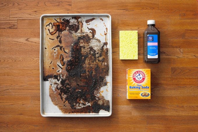 dirty baking sheet next to a sponge, hydrogen peroxide bottle, and baking soda box on wood butcher block background