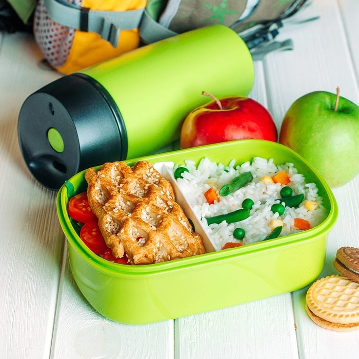 Lunch box filled with rice and sandwich near thermos, fresh apples and biscuits in front of kids school backpack on white wooden background
