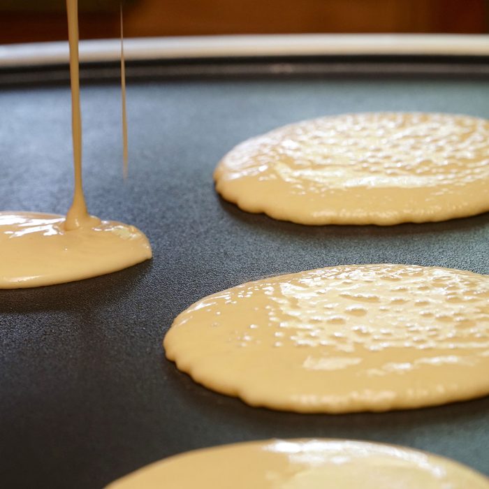 Pancake batter baking mix being poured from a bowl onto a hot electric griddle cooking delicious breakfast meal for a family.