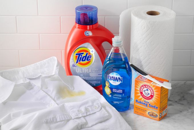 White Shirt Stained With oil next to strain removal supplies: tide detergent, dawn dish soap, baking soda, paper towels, and a toothbrush