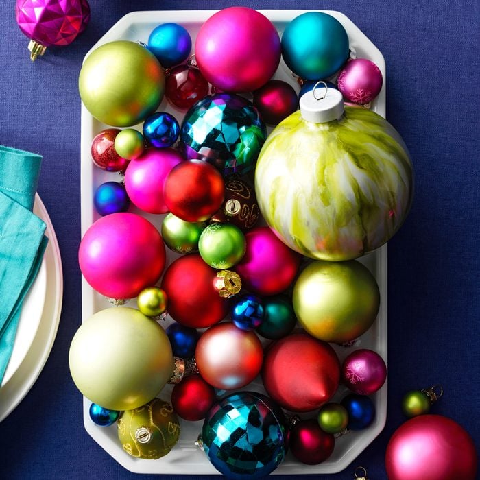 Overhead of Multicolored Assorted Ornaments Resting in a White tray on a linen background with ornaments scattered around a table place setting