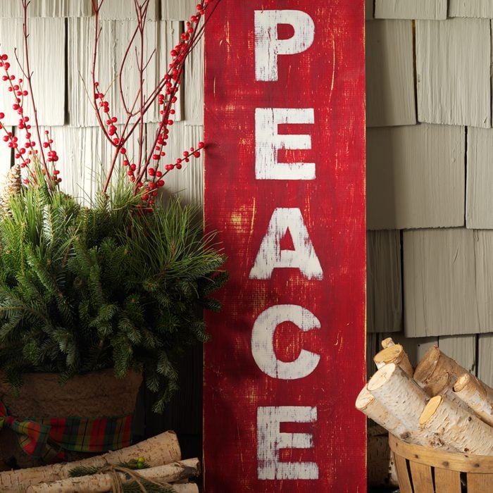 Christmas Porch Display with "Peace" Red sign against home siding with logs, assorted branches and Winter decorations displayed around the concrete pad
