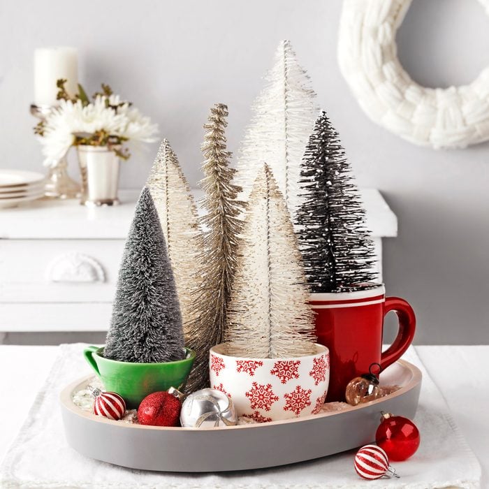 Christmas Tree Decorations Resting in Holiday Tea Cups on a Linen placemat against a bright white Christmas Holiday Decoration Setup