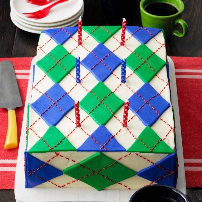 Wacky Argyle Cake; 3/4 camera angle; dark wood surface; large cake; candles; birthday candles; green mug; blue mug; coffee; coffee mugs; coffee mug; red plastic forks; plastic forks; frosting; fondant; red runner; red table cloth; white plate stack; plate stack; cake knife; cake server; white cutting board; rustic wood surface