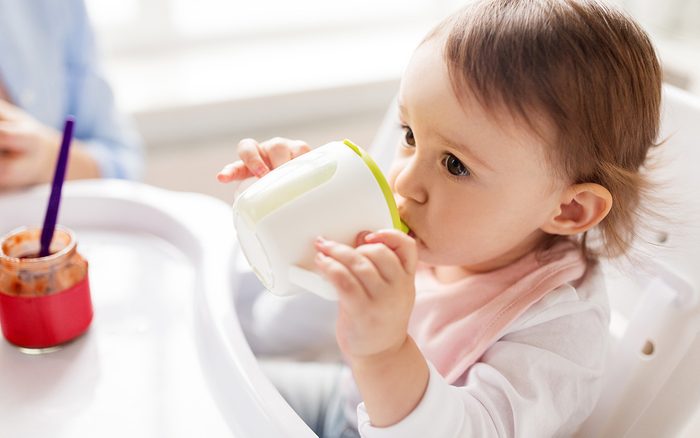 childhood and people concept - little baby drinking from spout cup sitting in highchair at home