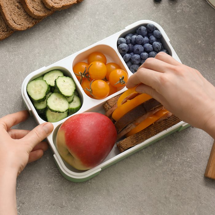 Mother putting food for schoolchild in lunch box on table