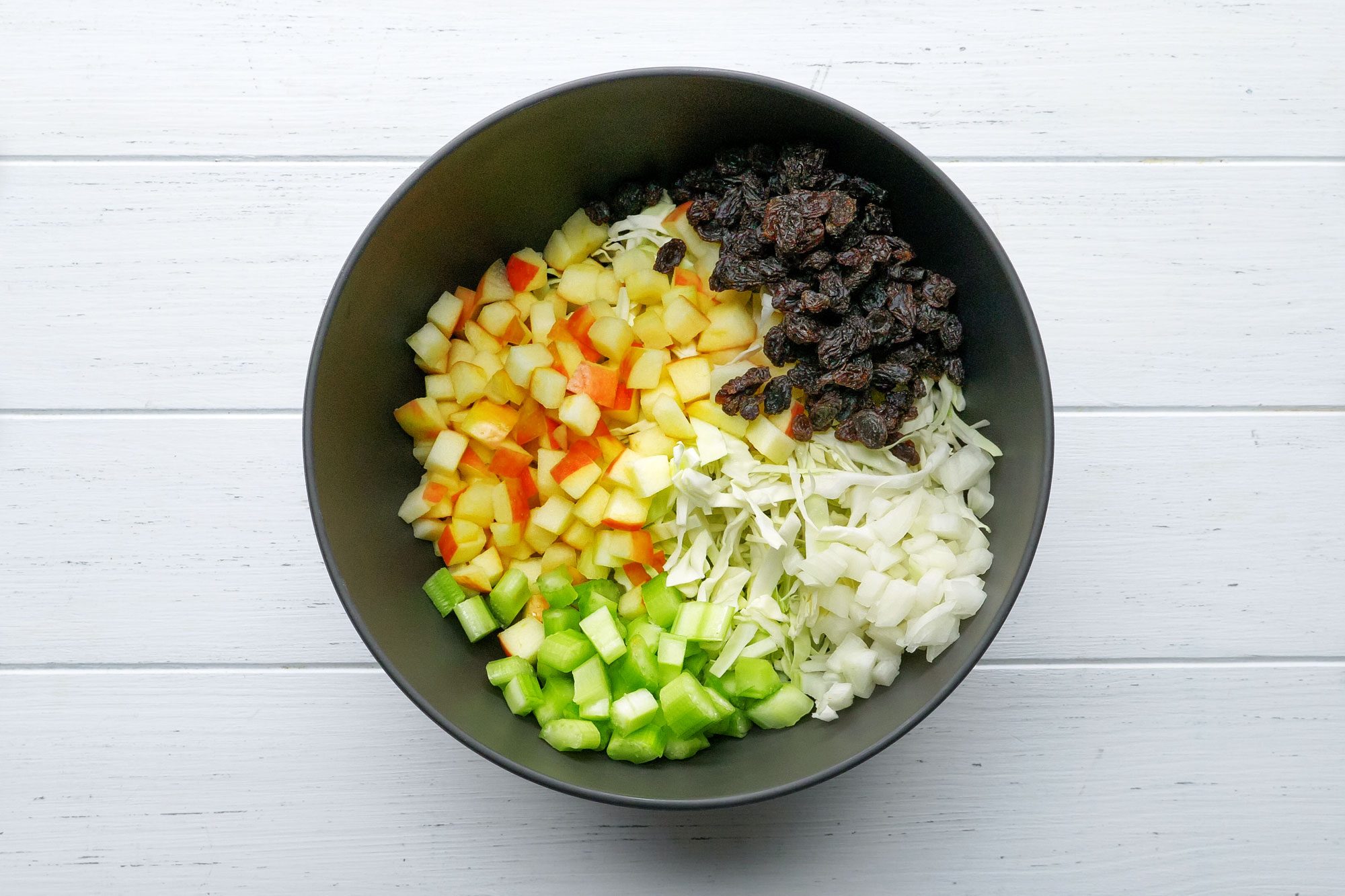 shredded cabbage, chopped apples, celery, raisins and onion in a large bowl on painted wooden surface
