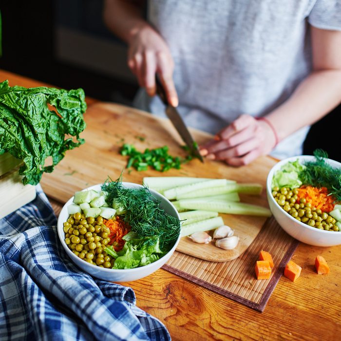 Bowl with green peas, cucumbers, carrots, lettuce and dill standing on a table on a background of hand chopping green onions with knife on wooden Board