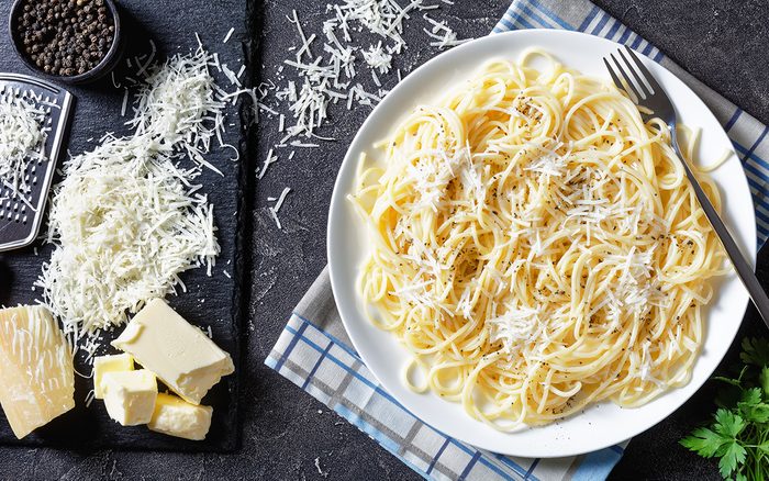 cacio e pepe, pasta mixed with grated pecorino cheese and dusted with freshly ground black pepper on a white plate with a fork. grated cheese, black pepper in a bowl on a concrete table