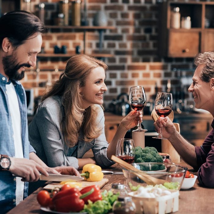 happy friends drinking wine and smiling each other while man cutting vegetables for salad