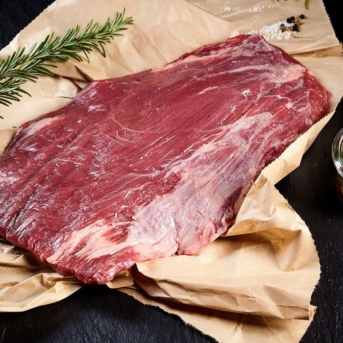 Portion of lean raw flank steak for roasting or grilling on crumpled brown paper in a rustic kitchen with a sprig of fresh rosemary and olive oil marinade