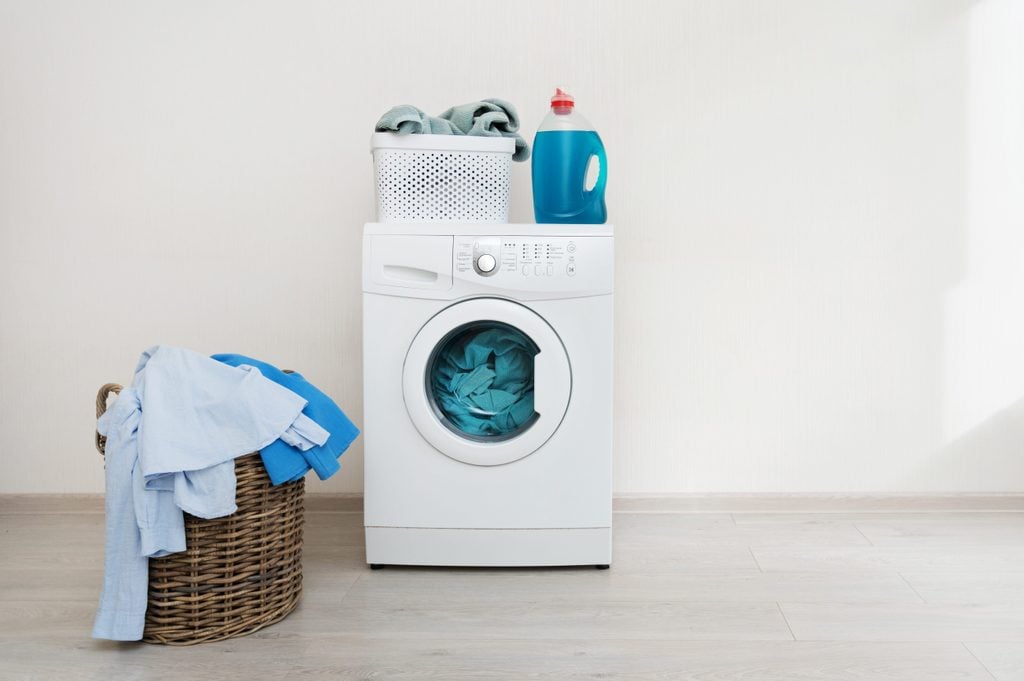 Photo of preparation to laundry process. White washing machine standing on laminate floor near wooden basket with clothes. Object isolated against wall inside bright light flat interior