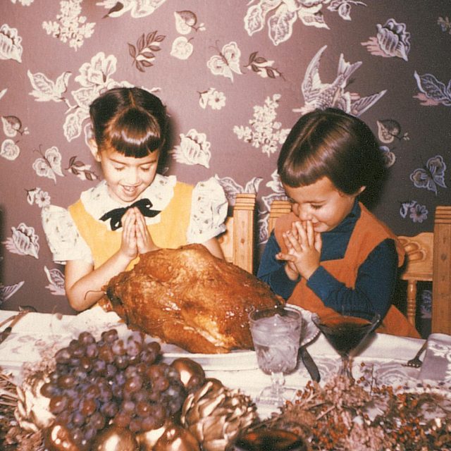 two 1950s sisters laugh while they prey for the turkey on the table