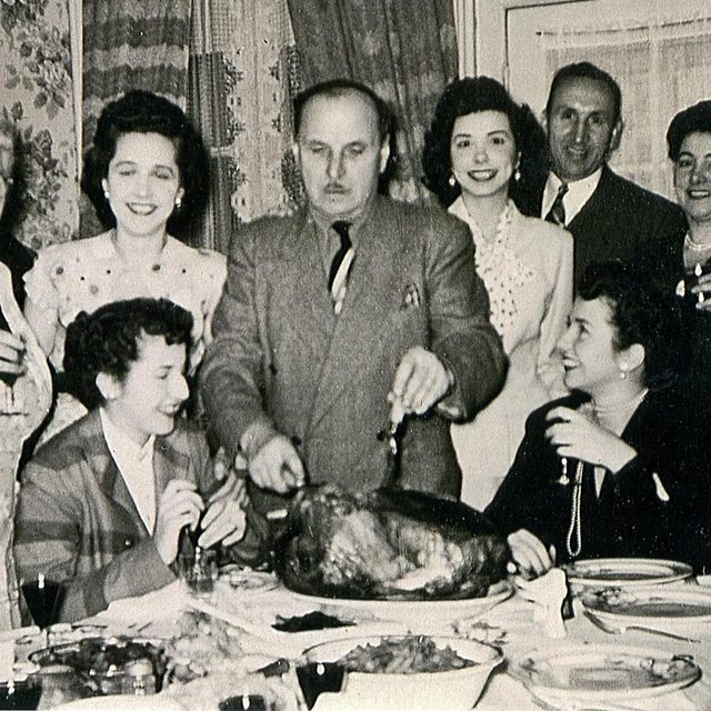 Man cuts turkey at the dinner table while young women smile at him and the camera