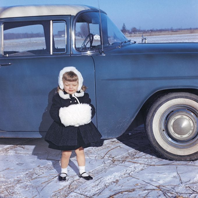 little girl stands in front of car dressed in a black winter outfit and hand warmers, 1960s