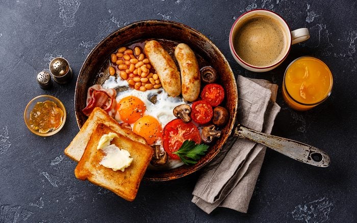 Full English breakfast in pan with fried eggs, sausages, bacon, beans, toasts and coffee on dark stone background