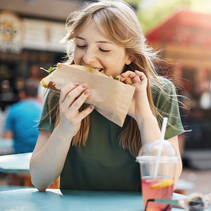 Hungry freckled blonde girl eating taco on a food court on a sunny summer day in park.