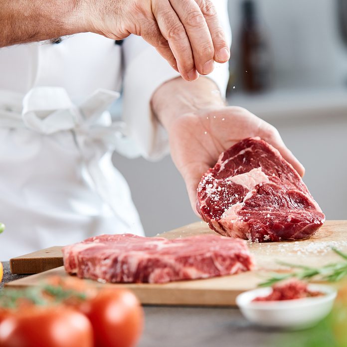 Chef sprinkling cooking salt onto a raw ribeye steak while preparing dinner with fresh salad ingredients and herbs on the counter; Shutterstock ID 1181786554; Job (TFH, TOH, RD, BNB, CWM, CM): Taste of Home