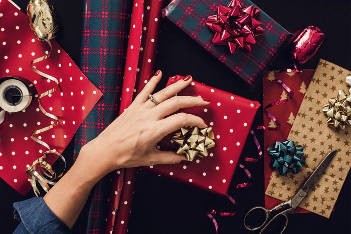 overhead view of a hand adding a bow to a christmas present. gift wrapping paper, bows, ribbon, and a pair of scissors are scattered around the frame