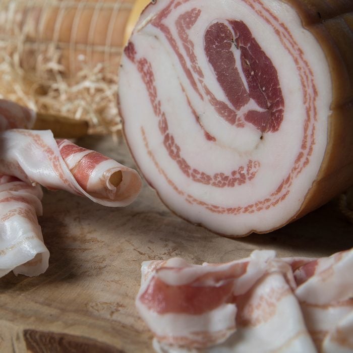 closeup of a wooden chop board with slices of italian pancetta made in Piacenza, north italy. Slices are twisted around bread sticks called grissini in Italian.