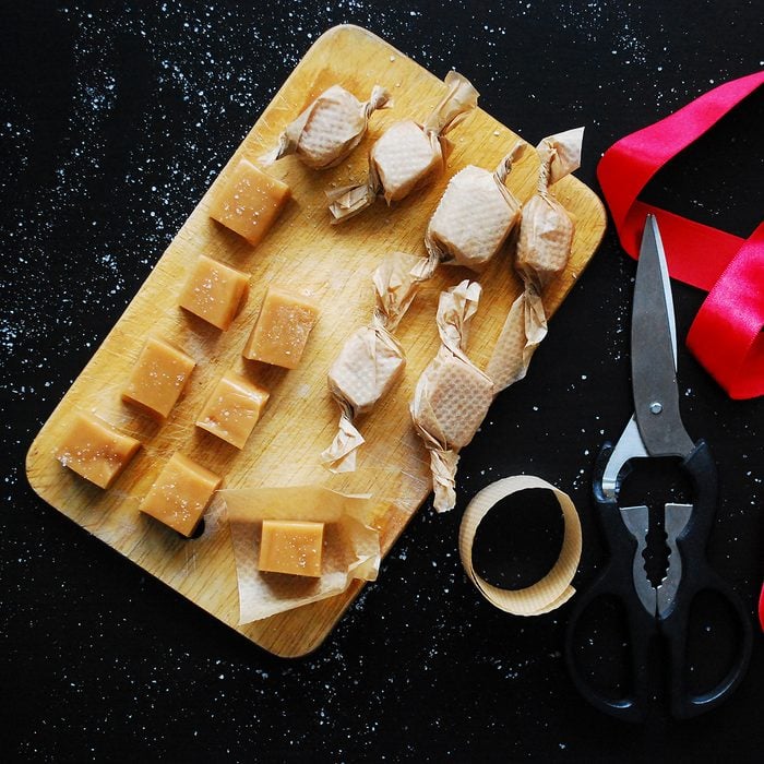 Homemade soft caramel candies wrapped in parchment paper with red ribbon and scissors on black background. Top view.