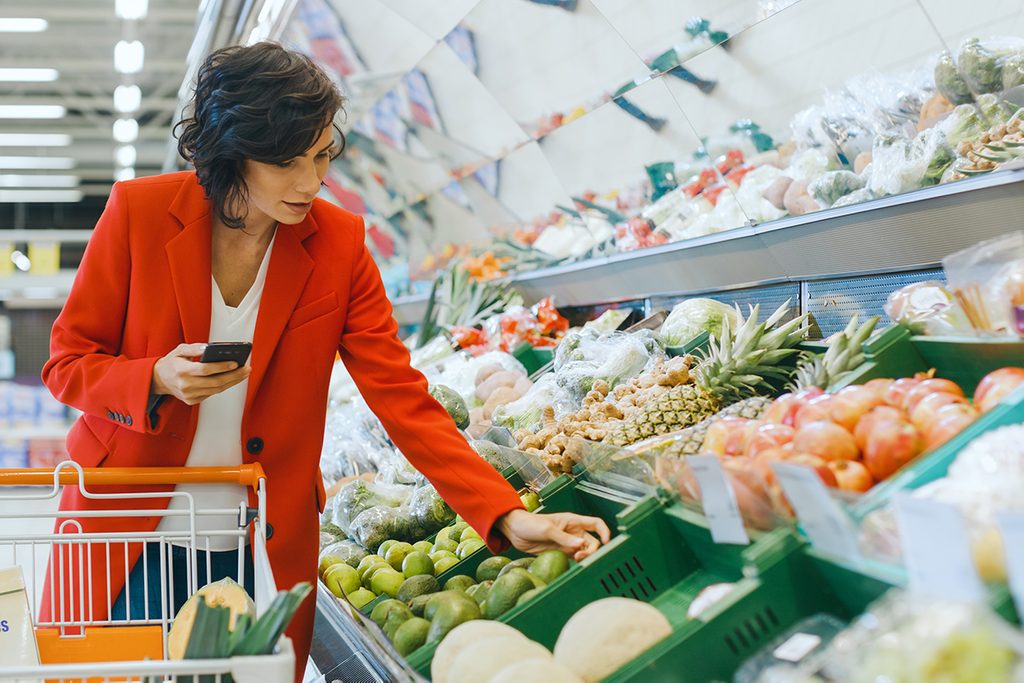 At the Supermarket: Beautiful Young Woman Walks Through Fresh Produce Section, Chooses Vegetables and Places them in Her Shopping Cart. Customer Uses Smartphone while Shopping for Fruits; Shutterstock ID 1275256033; Job (TFH, TOH, RD, BNB, CWM, CM): Taste of Home