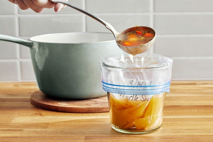 Hand using a ladle to transfer soup into a plastic freezer bag from a pot in a kitchen setting