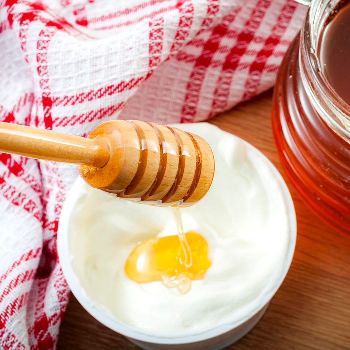 Honey dripping from dipper in a yogurt cup next to a glass jar of honey and a tea towel in a rustic kitchen