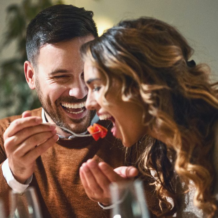 Closeup of mid 20's couple having fun during dinner party. The guy is feeding his girls with some chopped fruit, both laughing.