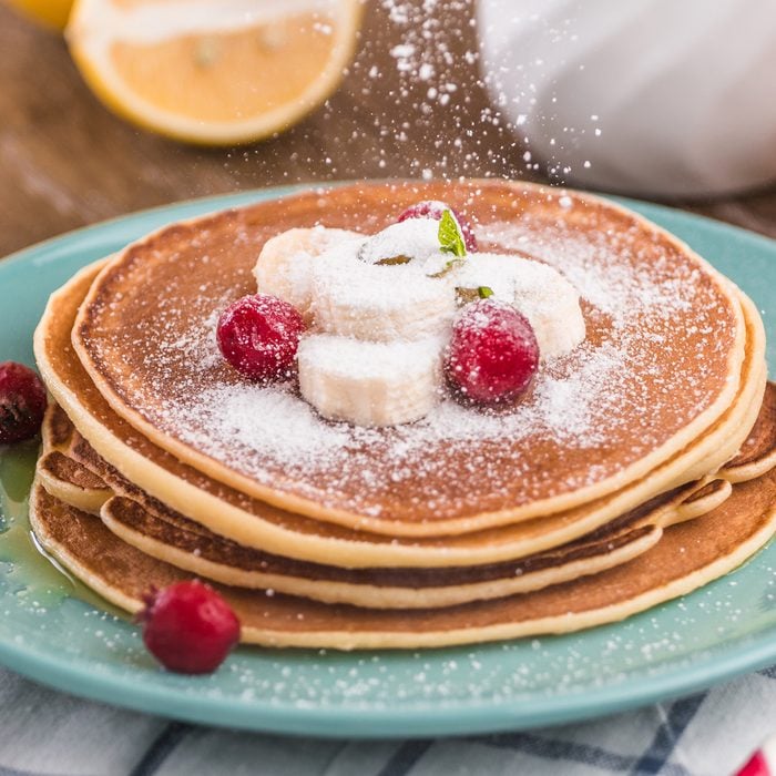 Pancake stack covered with honey, powdered sugar, banana and cranberries. On a wooden background in a blue plate