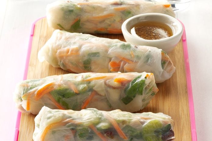 Four homemade spring rolls arranged on a cutting board with a bowl of dipping sauce.