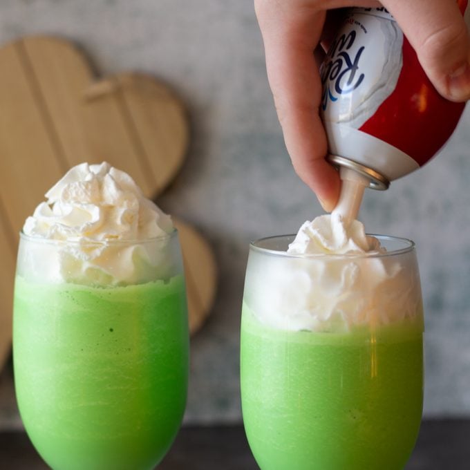 Two glasses with green milkshakes and whipped cream being added to tops.