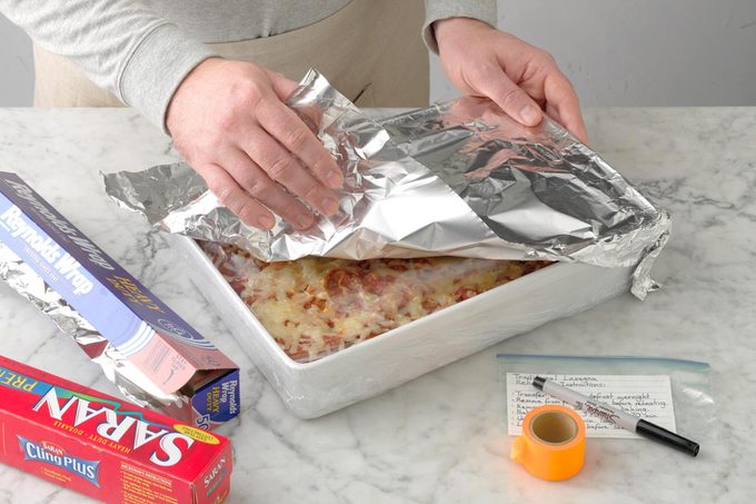 how to freeze lasagna; marble surface; how to; plastic wrap; foil, marker; colored washi tape; hands