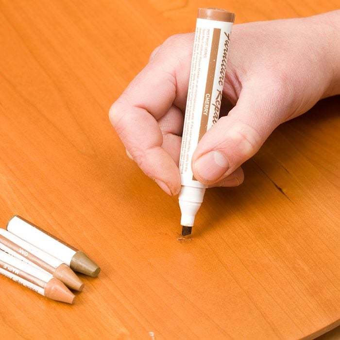 A worker is using a furniture repair marker to color fix a scratched cherry table surface. Three repair sticks rest to the left.