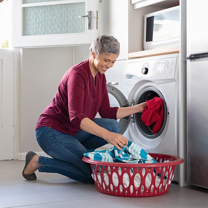 Happy senior woman loading dirty clothes in washing machine. Smiling mature woman sitting on floor putting clothed in washing machine from laundry basket. Housework.
