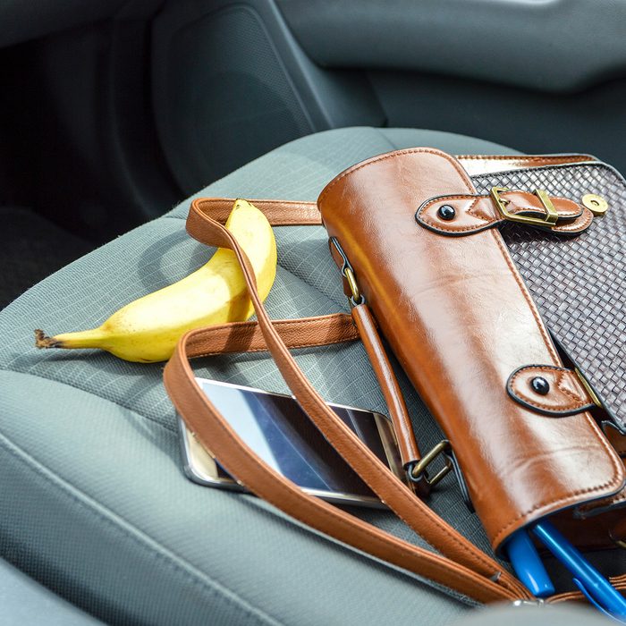 This is an image of a brown purse on the passenger seat of a car with contents spilling out. There is a pen, makeup item, phone and banana laid out around the purse. The car interior is grey.